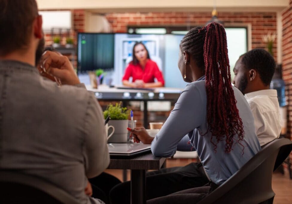Black woman and coworkers sit at table, attentively listening to client on video call displayed on monitor. Selective focus on african american lady looking at screen showing virtual call with employer